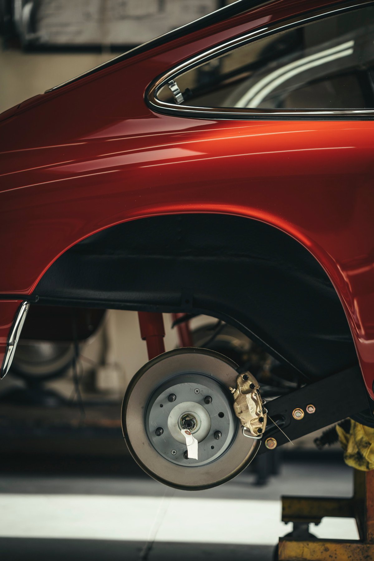 Vintage red Porsche 911 rear quarter, detailed chrome trim, exposed brake rotor, and gold caliper during restoration.