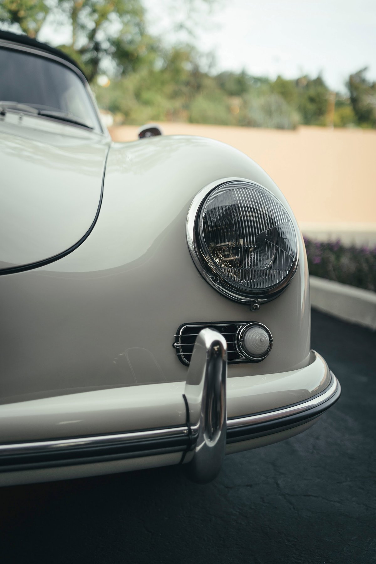 Close-up of a classic ivory Porsche 356 Cabriolet's front. Iconic round headlight, chrome trim, bumperette, and signal light.