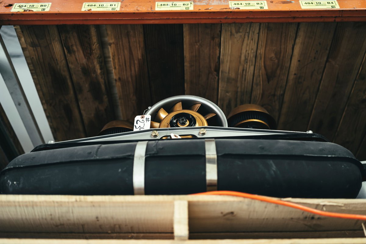 Porsche 911 air-cooled engine components: gold cooling fan, black fuel tank with straps, and dual air filter housings.