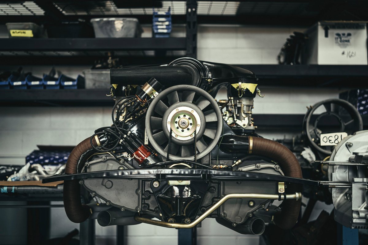 Restored Porsche 911 air-cooled flat-six engine with its iconic cooling fan and brown wrapped exhaust in a workshop setting.
