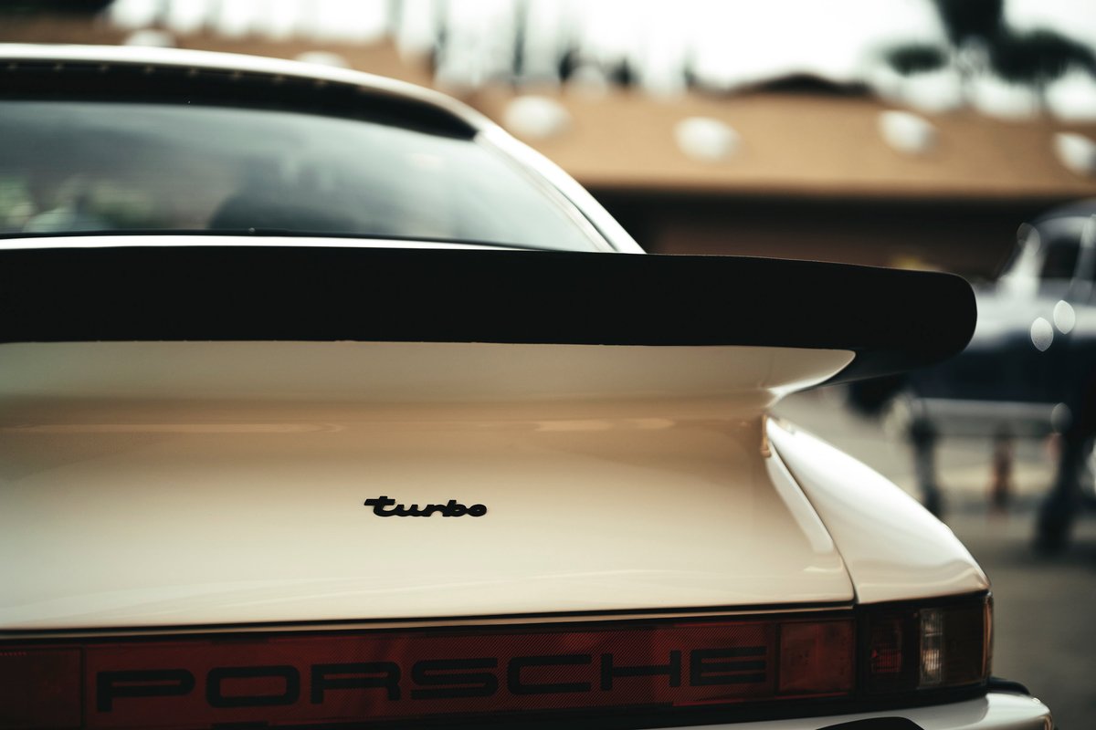 White Porsche 930 Turbo rear with iconic black "whale tail" spoiler, "turbo" decklid badging, and PORSCHE taillight script.
