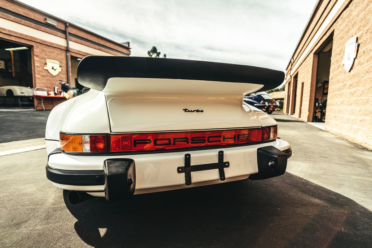 White Porsche 930 Turbo with iconic black whale tail spoiler, 'PORSCHE' script rear reflector panel, and classic bumperettes.