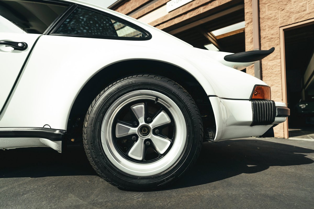 White Porsche 911 G-series rear quarter, showcasing classic Fuchs rims, 'whale tail' spoiler, and black impact bumper.