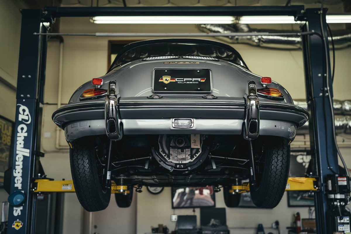 Silver Porsche 356 coupe on a service lift, rear view. Showcases vintage chrome bumper, classic taillights, & exposed transaxle.