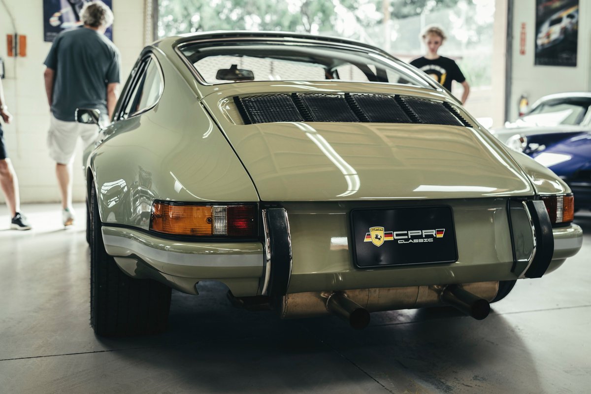 Vintage sage green Porsche 911 rear, featuring iconic engine lid grilles, chrome trim, bumperettes, and dual exhaust.