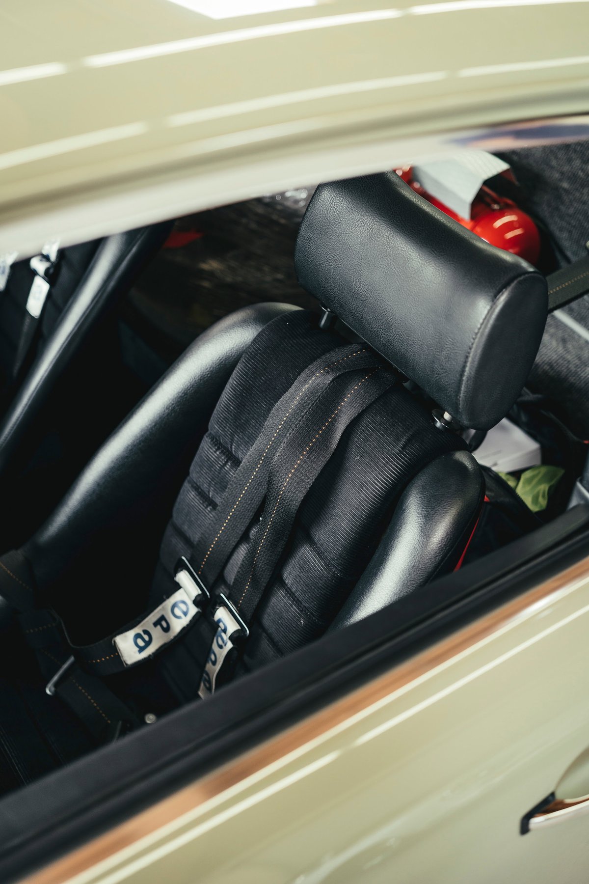 Cream/beige air-cooled Porsche 911 interior. Black OMP racing bucket seat, multi-point harness, roll cage, and fire extinguisher.