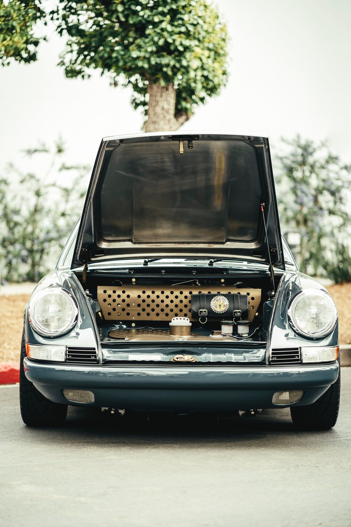 Front view of a Slate Grey Porsche 911. Its frunk is open, revealing custom perforated brass trim and a leather tool roll.
