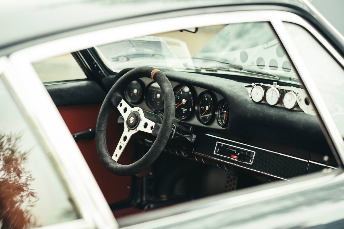 Interior of a classic air-cooled Porsche 911, showcasing its Alcantara steering wheel, five-gauge cluster, red trim, and rally timers.