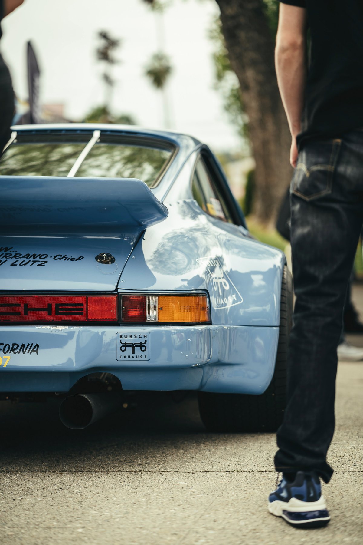 Rear view of a light blue widebody Porsche 911 featuring a large whale tail spoiler, custom decals, and Bursch exhaust.