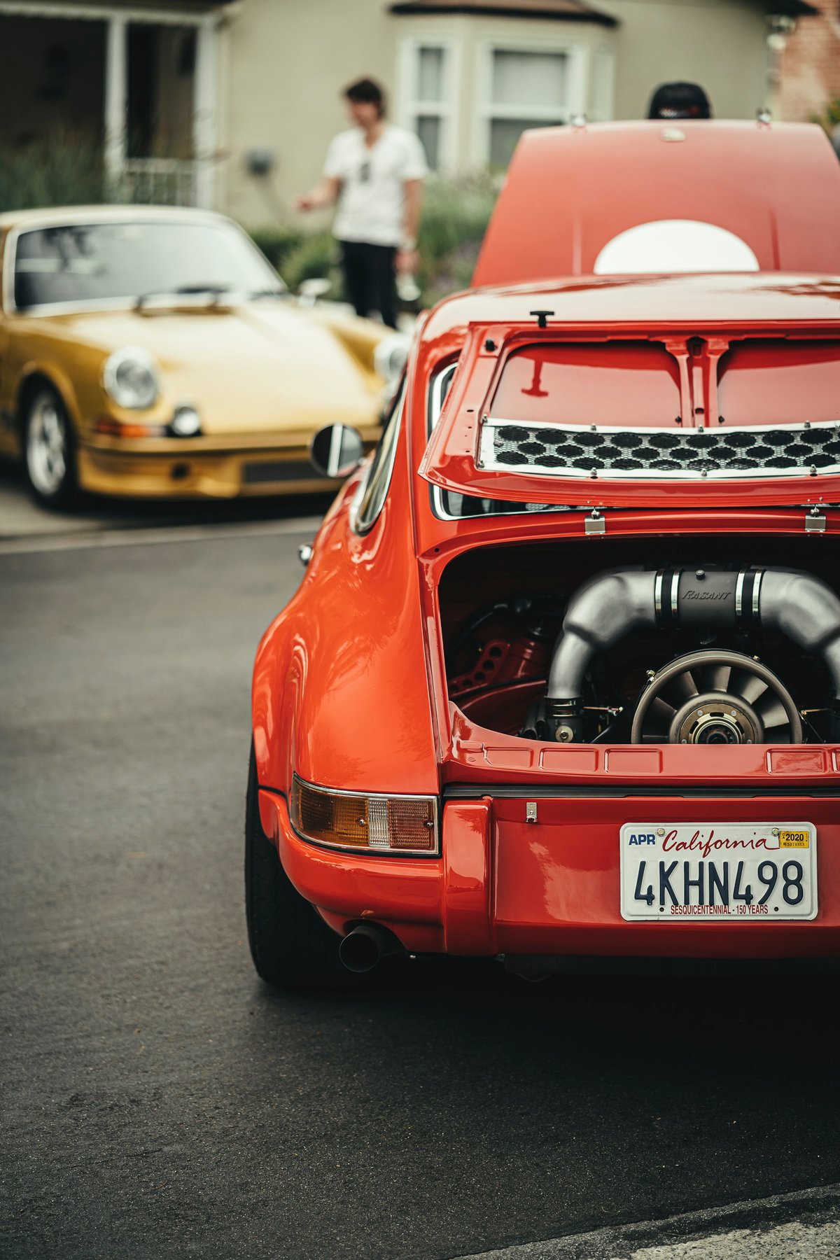 Vibrant red custom air-cooled Porsche 911's rear engine bay showcased, revealing intricate intake system and engine fan. Gold 911 nearby.