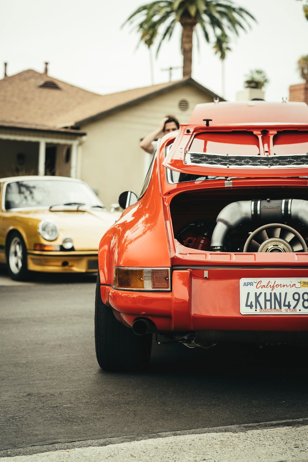 Rear view of a vibrant orange air-cooled Porsche 911 with its engine bay open, showcasing the detailed flat-six engine.