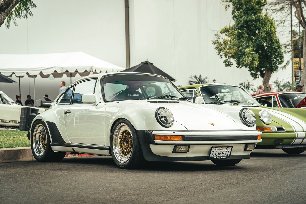 White Porsche 930 Turbo with wide-body fenders, black trim, and gold multi-spoke rims at a Los Angeles car show.