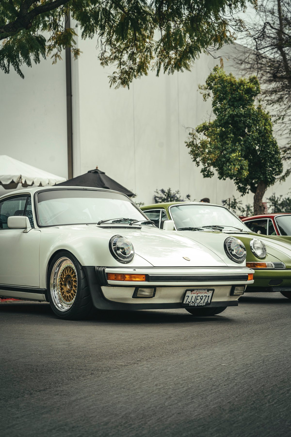 White Porsche 911 G-series coupe with gold multi-piece mesh rims and black bumper trim, parked with other classic 911s.