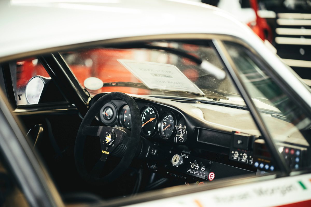 White air-cooled Porsche 911 race interior: black MOMO suede wheel, classic gauges, minimal competition dash, visible roll cage.