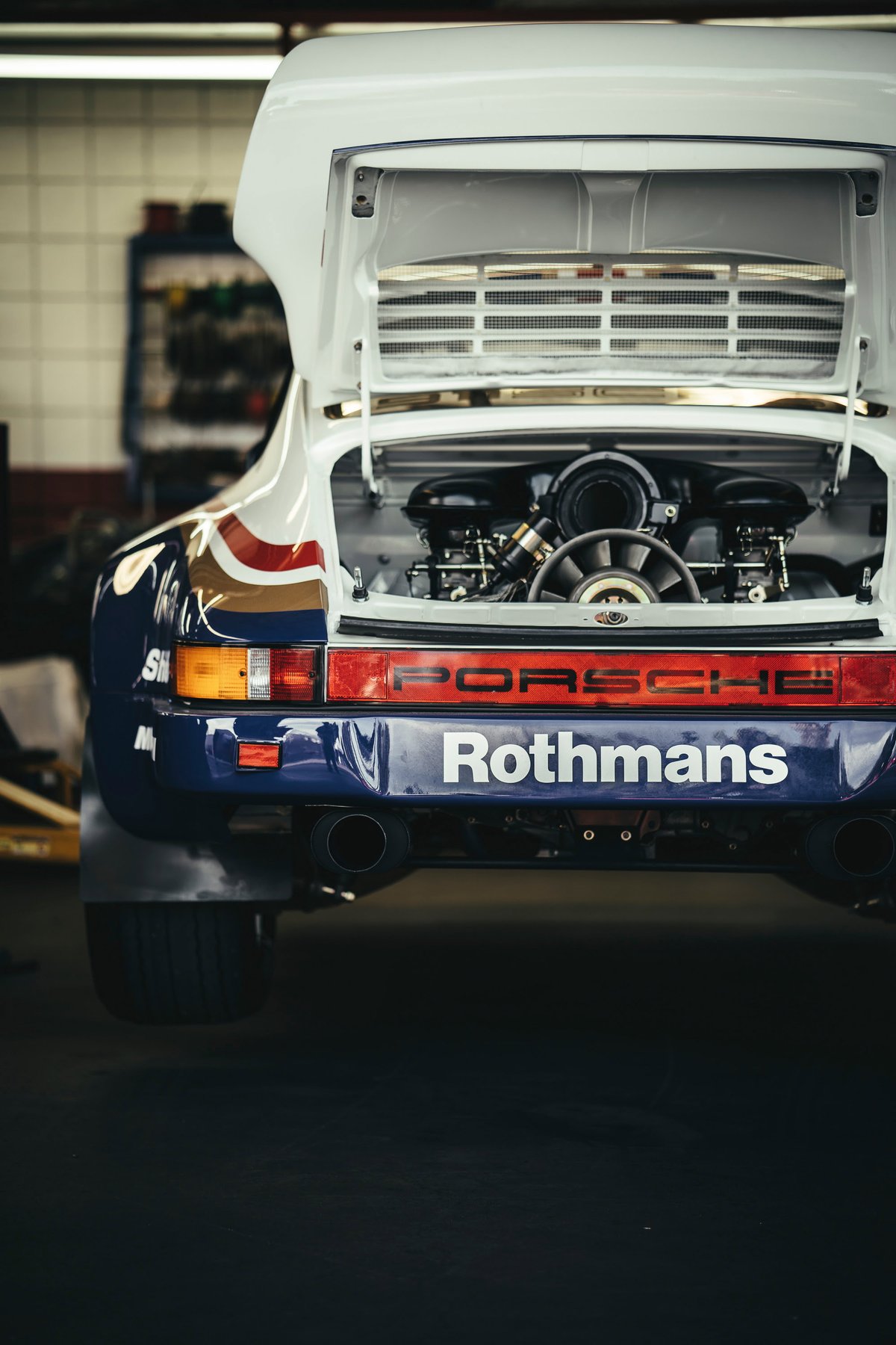 Classic Porsche 911 rally car, white with Rothmans livery. Open decklid exposes its flat-six engine. Rear view shows mud flaps & dual exhausts.