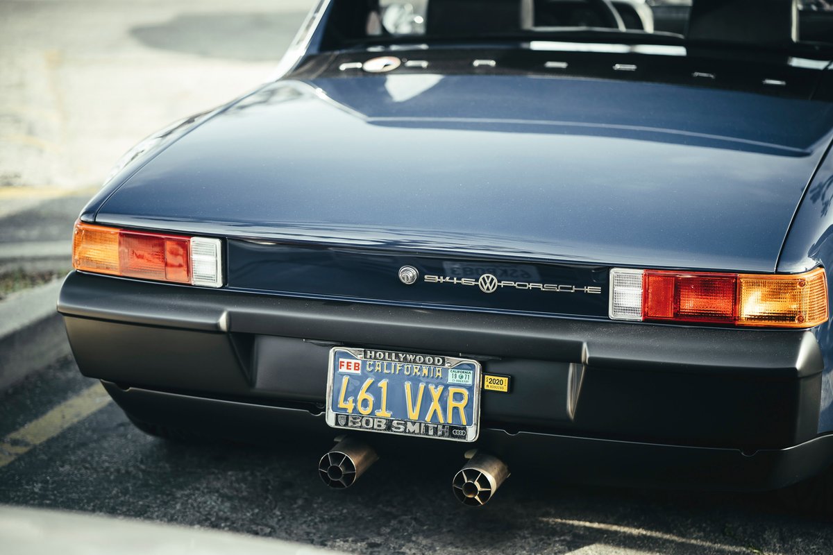 Rear detail of a classic dark blue Porsche 914. Features segmented taillights, black bumper, VW-Porsche badging, and custom twin exhaust tips.