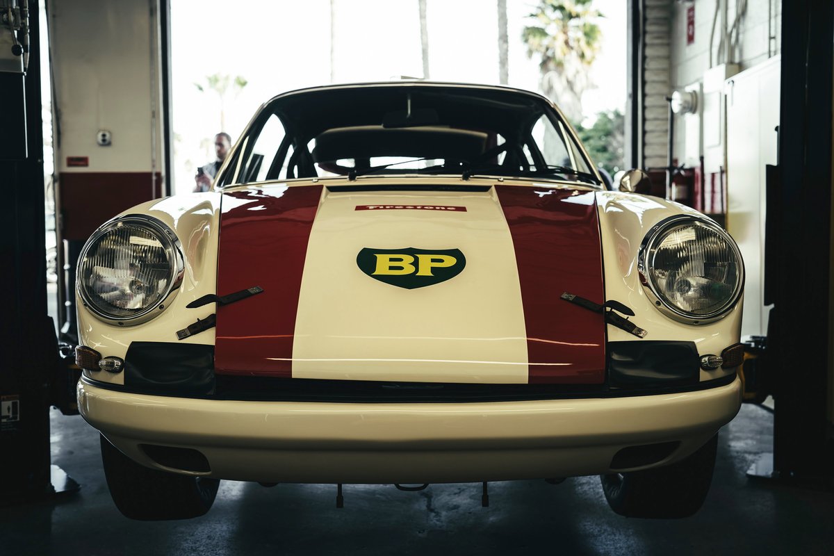 Vintage air-cooled Porsche 911 race car, white with red stripe & BP livery. Front fascia, classic headlights, and hood pins.