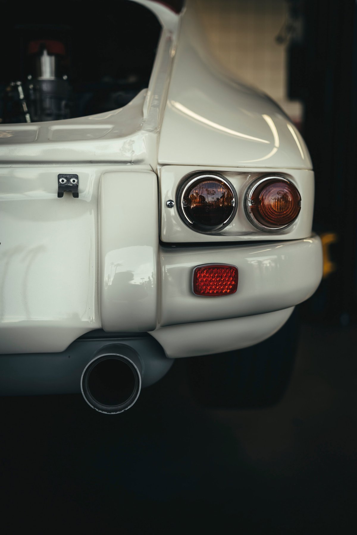 Rear detail of a light ivory classic Porsche 911 with round taillights, amber turn signal, red reflector, and single exhaust.