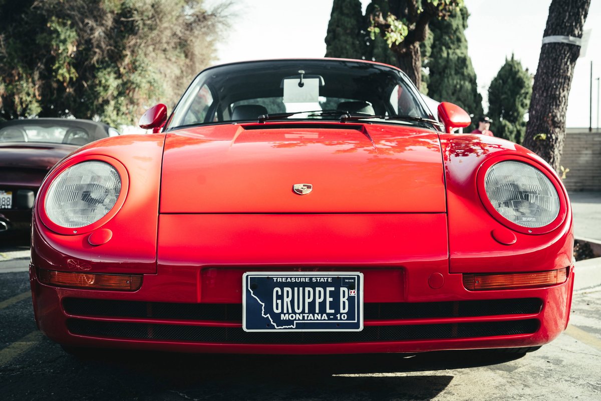 Bright red Porsche 959 hypercar front view, highlighting its distinctive headlights, fascia, and custom "Gruppe B" plate.
