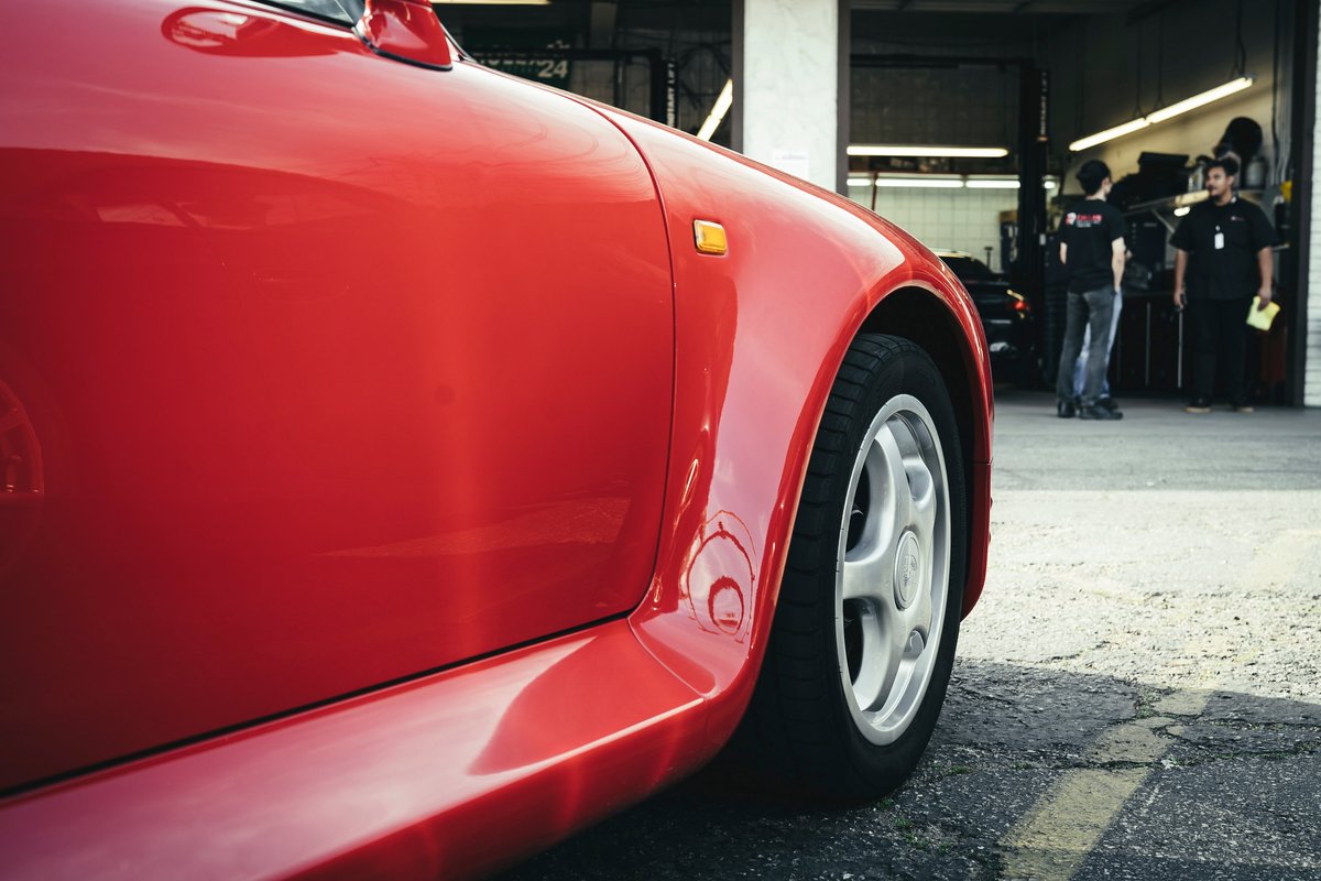 Close-up of a vibrant red Honda S2000's front fender, amber side marker, and silver 5-spoke AP1 OEM rims in a garage.