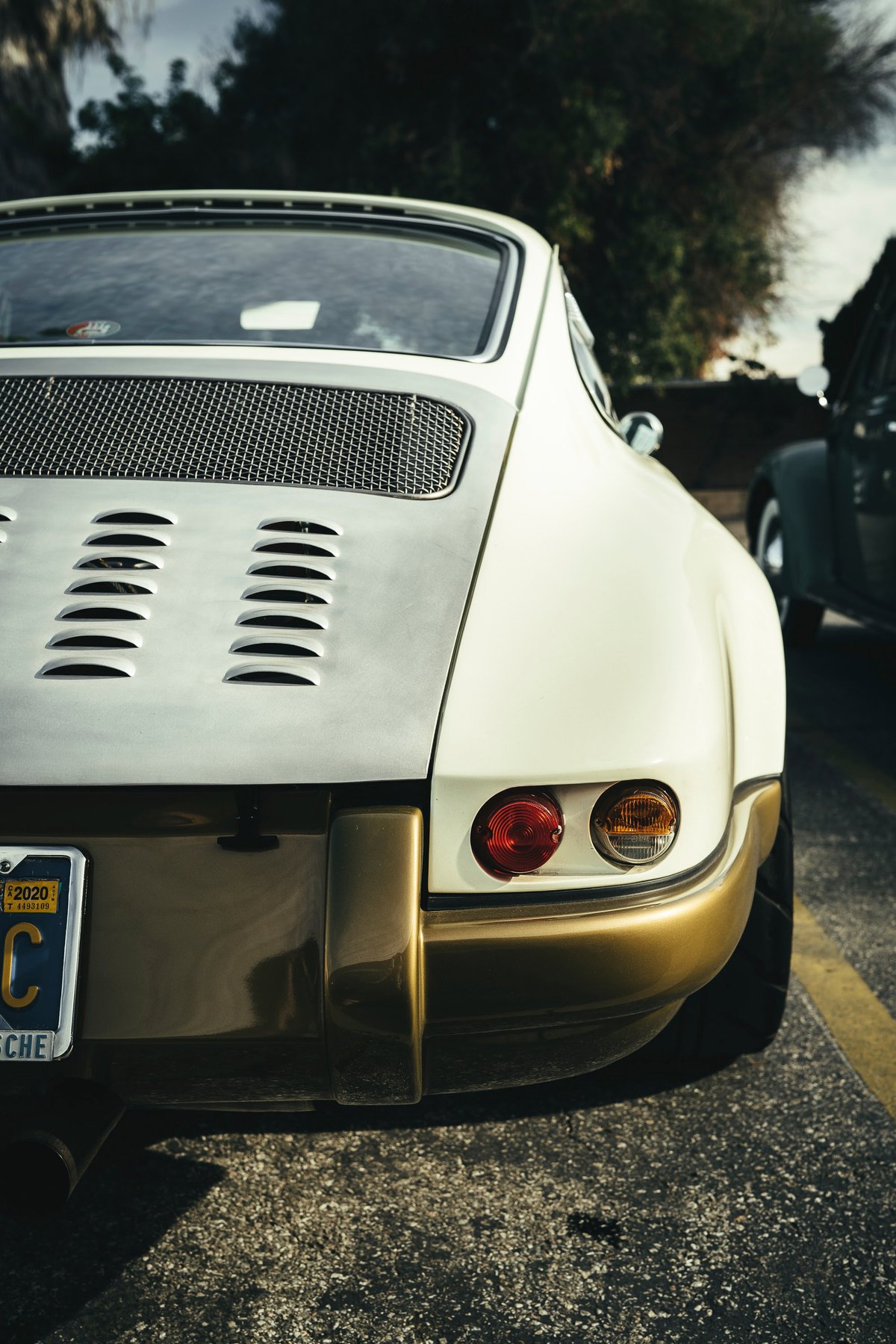 Rear view of a classic two-tone Porsche 911, off-white with bronze lower bumper & trim. Engine lid features a mesh grille and horizontal louvers.
