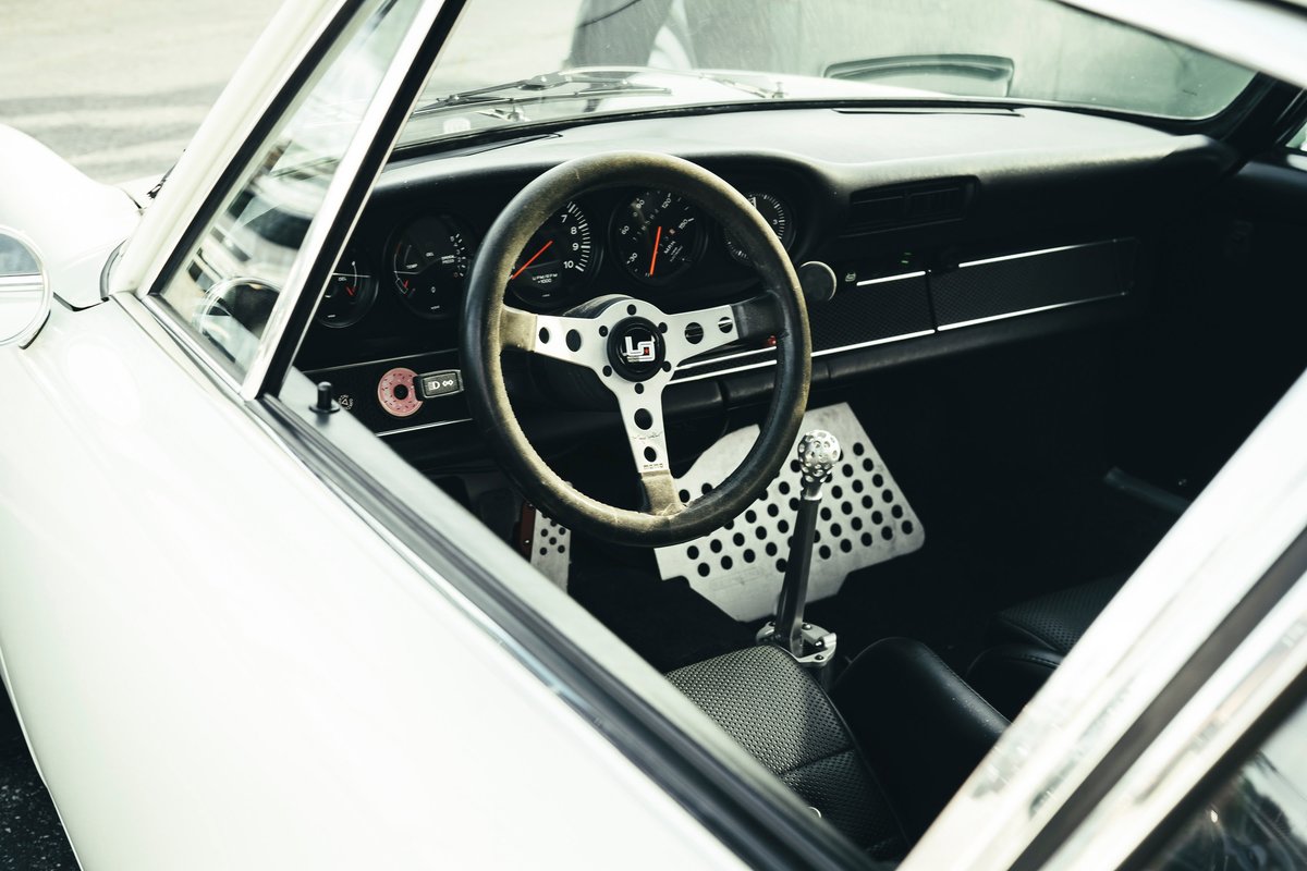 Classic white Porsche 911 interior: aftermarket racing steering wheel, perforated aluminum pedals, and manual shifter.