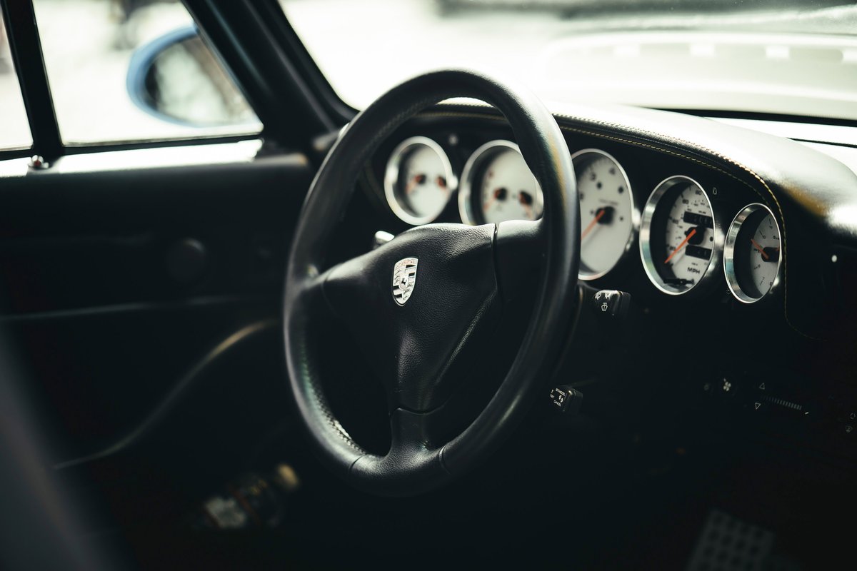 Classic Porsche 911 interior: black leather steering wheel with crest, white-faced five-dial instrument cluster, and dark dash trim.