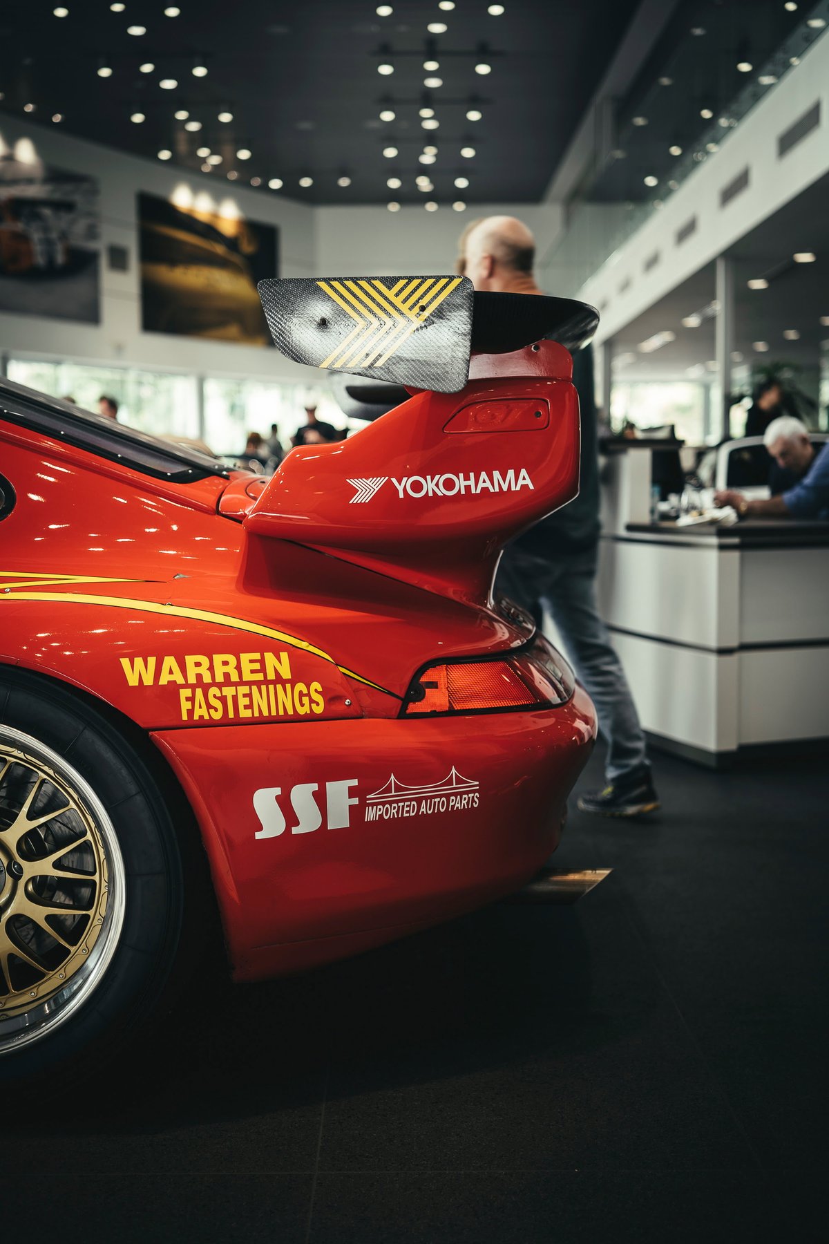 Red Porsche 993 race car with a carbon rear wing, Yokohama livery, and gold multi-spoke rims at a Los Angeles show.