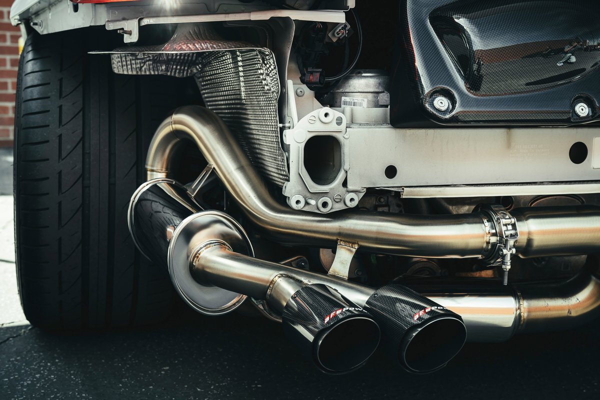 White performance car rear quarter: Akrapovič polished stainless steel exhaust, dual carbon fiber tips, exposed chassis, wide tire.