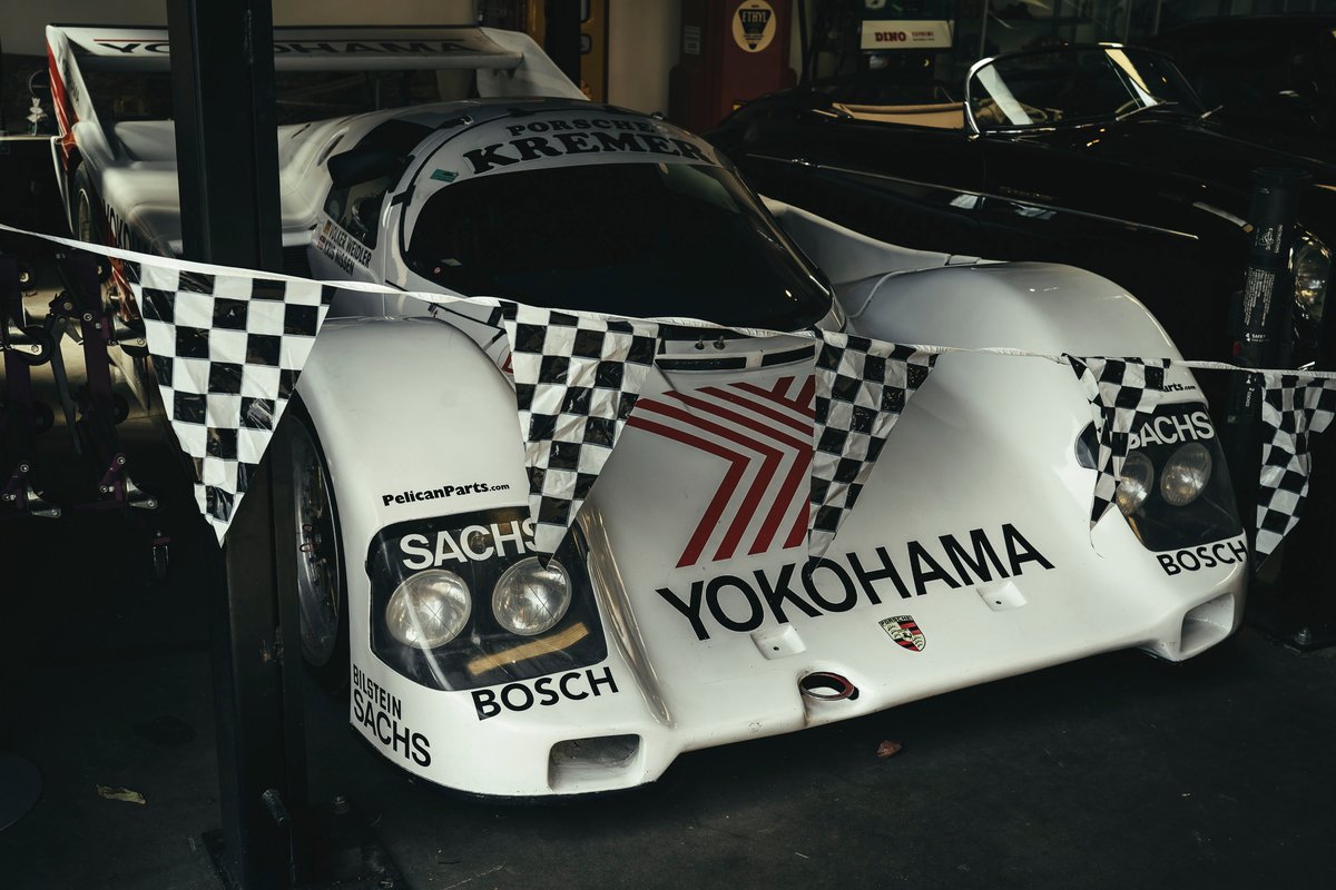 Iconic white Porsche 962C Kremer Group C prototype, featuring YOKOHAMA/SACHS livery and exposed racing rims, draped with flags.