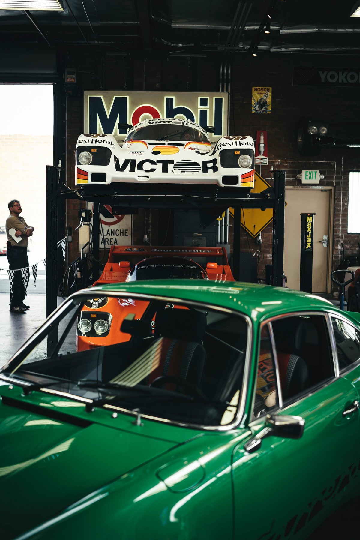 Classic green Porsche 911 coupe. A white Porsche 962 race car with Victor livery is on a lift, an orange Porsche 935 below.