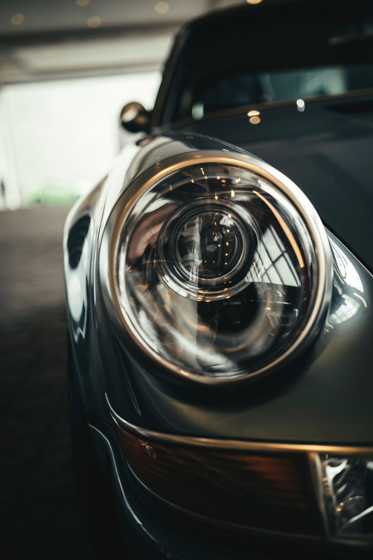 Close-up of a classic Slate Grey Porsche 911 headlight, highlighting its iconic round design, polished chrome bezel, and amber turn signal.