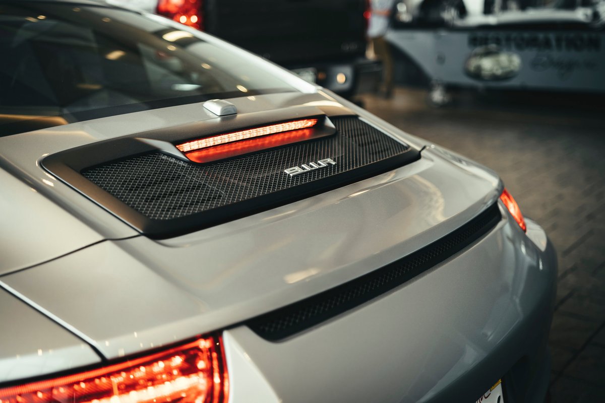 Silver Porsche 992 911 rear view, showcasing its illuminated LED taillight strip, engine cover's mesh grille, and 911 badge.