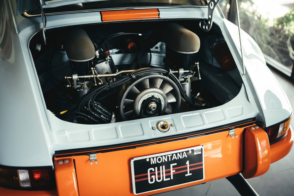 Exposed engine bay of a classic Porsche 911 in iconic Gulf livery, showcasing its air-cooled flat-six with fan and carbs.