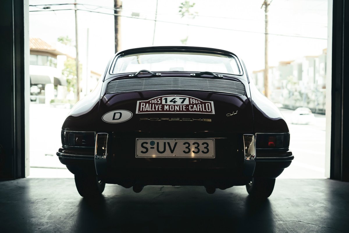 Rear view of a dark red 1965 Porsche 911, showcasing Monte Carlo Rallye livery, chrome bumperettes, and German plates.