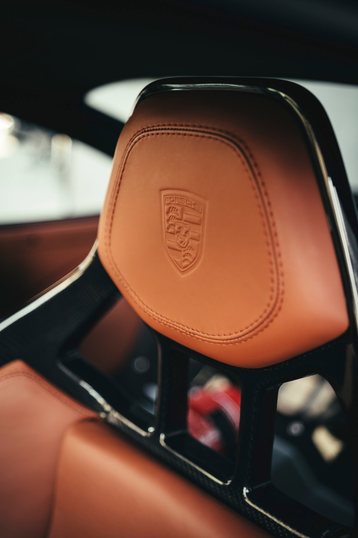 Porsche performance bucket seat headrest in rich tan leather, embossed crest, and visible carbon fiber frame details, premium interior.