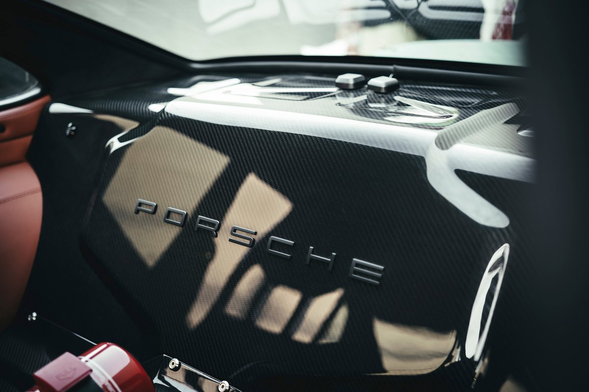 Porsche 911 interior features carbon fiber rear deck with metallic PORSCHE emblem, tan leather trim, and motorsport fire extinguisher.