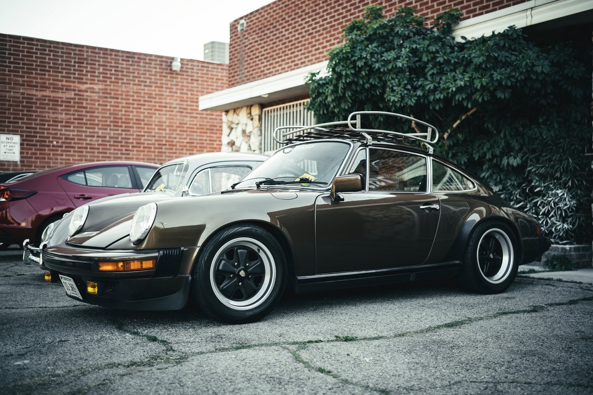 Vintage dark brown Porsche 911 with Fuchs-style rims, widebody flares, and period roof rack at a Los Angeles car show.