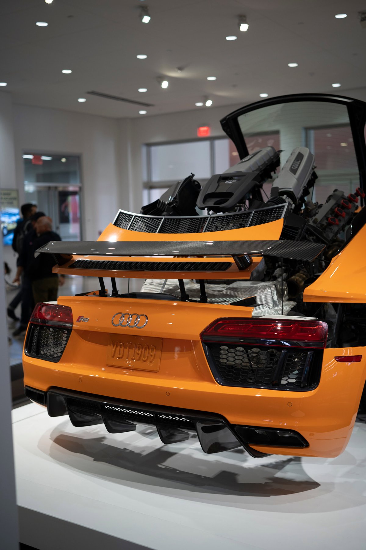 Orange Audi R8 V10 rear on display. Exposed mid-engine, carbon fiber wing, and aggressive black rear diffuser. Auto show exhibit.