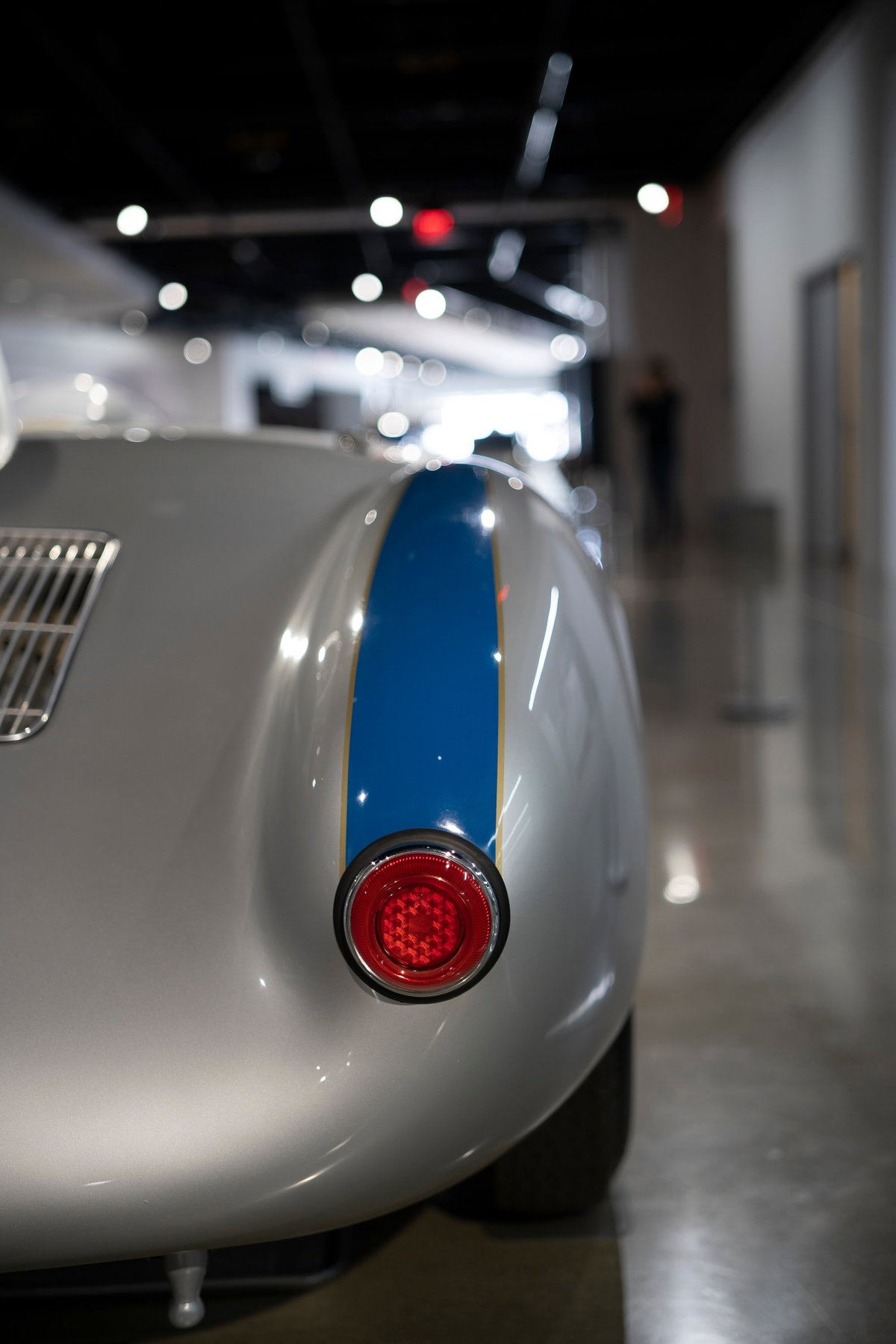 Close-up rear of a silver Shelby Cobra Daytona Coupe. Features iconic blue racing livery with gold pinstripes, classic round taillight, and rear vent.