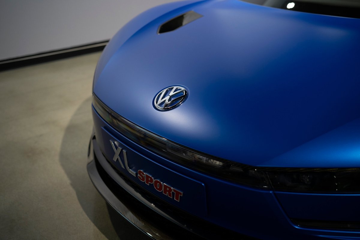 Close-up: Matte blue Volkswagen XL Sport front, showing VW emblem, "XL SPORT" badging, sleek headlights & carbon fiber trim.