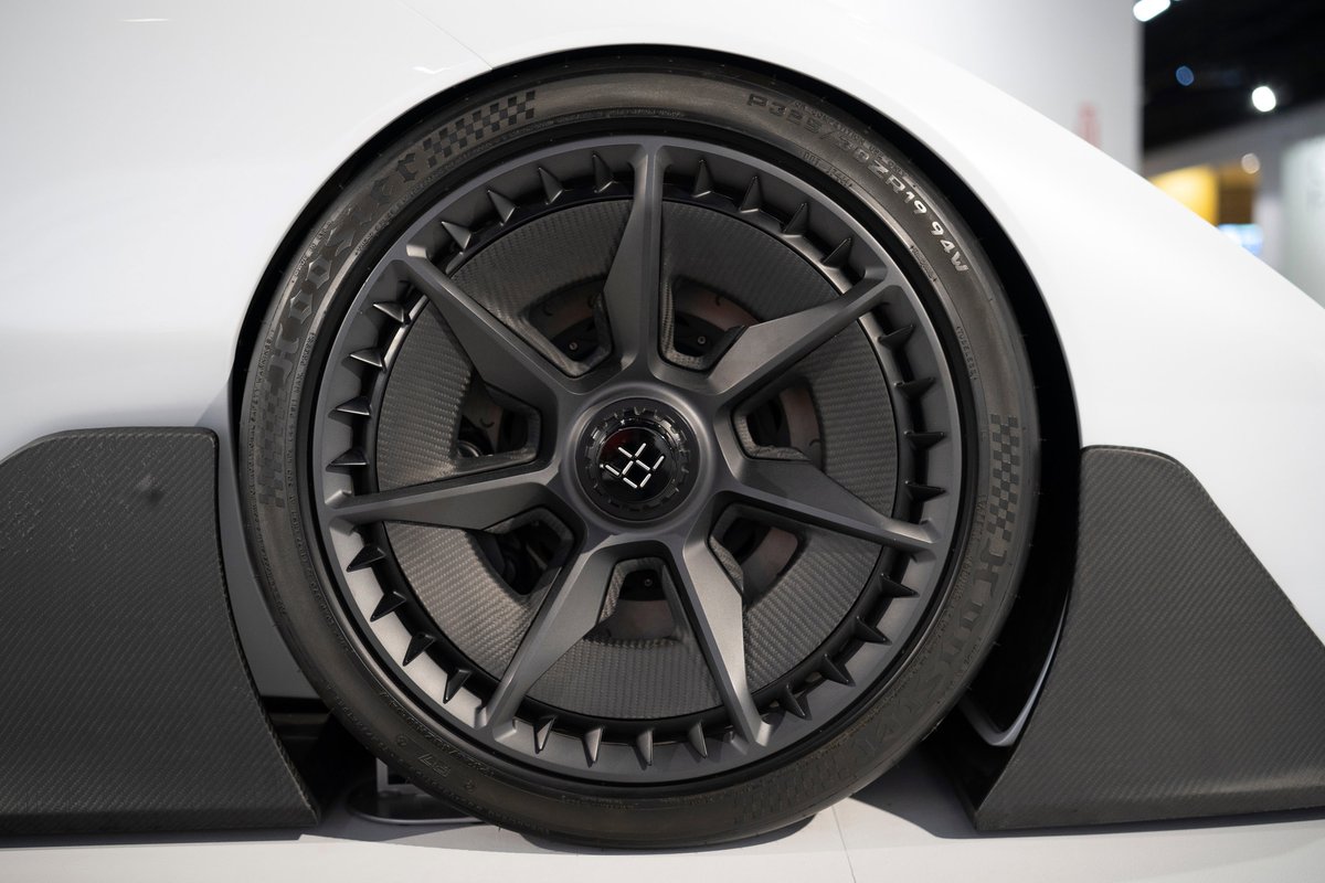 Close-up of a white Deus Vayanne electric hypercar's front wheel. Features dark multi-spoke rims, performance tires, & carbon fiber aero trim.