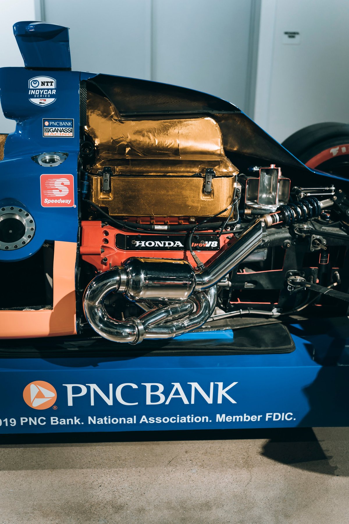 Exposed Honda Indy V8 engine with polished chrome exhaust headers and gold heat shield in a blue NTT IndyCar race car.