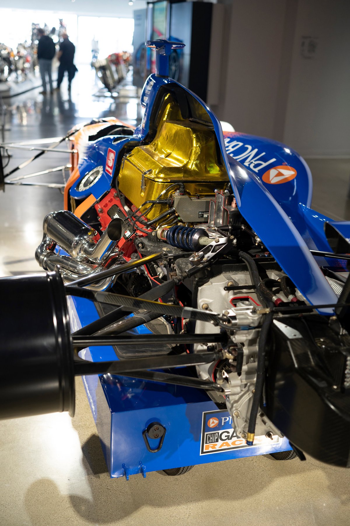 Exposed Dallara IndyCar engine: blue Chip Ganassi Racing livery, gold heat-wrapped components, chrome headers, carbon fiber suspension.