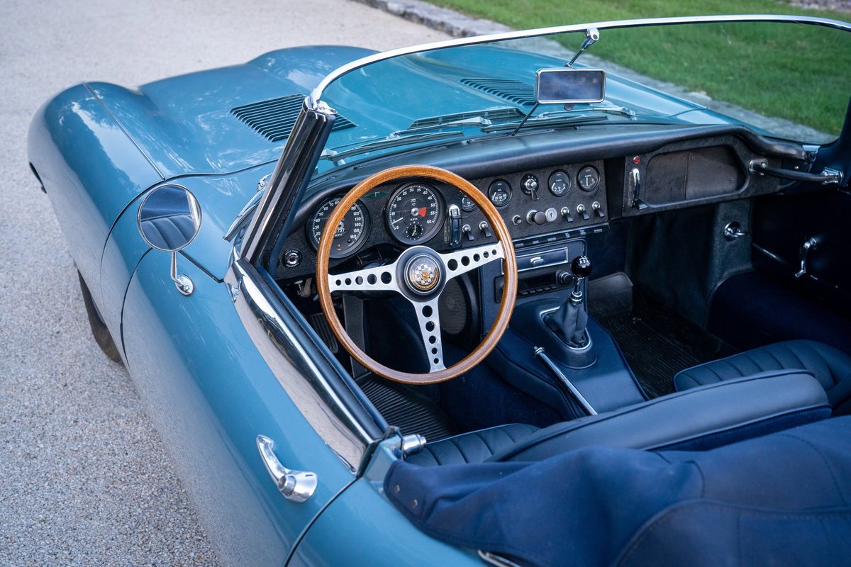 Interior of an opalescent silver-blue 1966 Jaguar XKE roadster with wood-rimmed steering wheel and blue leather seats.