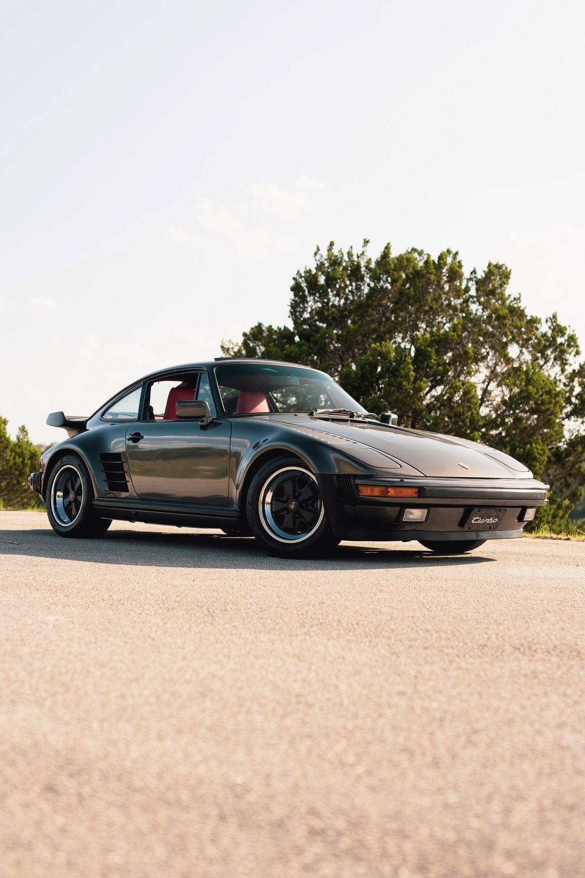 A black 1989 Porsche 930 Turbo Flachbau with red interior and black Fuchs rims.