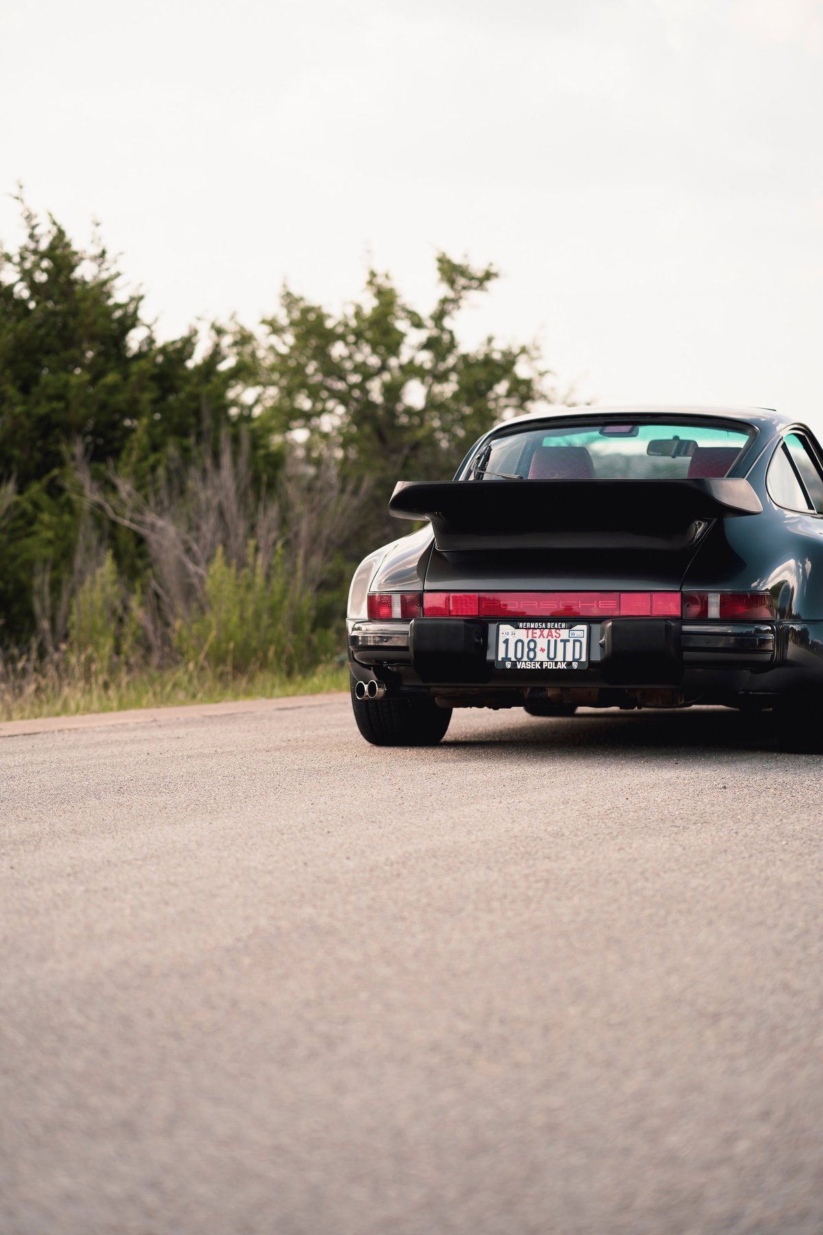 Black 1989 Porsche 930 Turbo Flachbau rear view with whale tail spoiler, dual exhaust, and aftermarket rims.