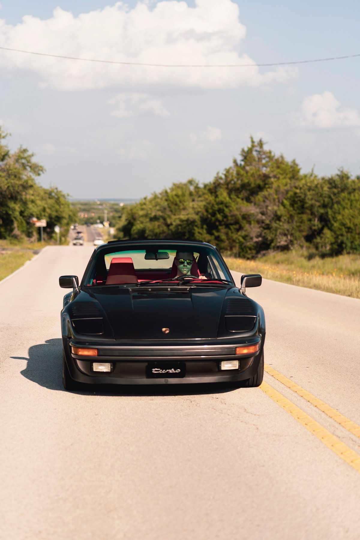 Black 1989 Porsche 930 Turbo Flachbau with red interior driving on a rural road.