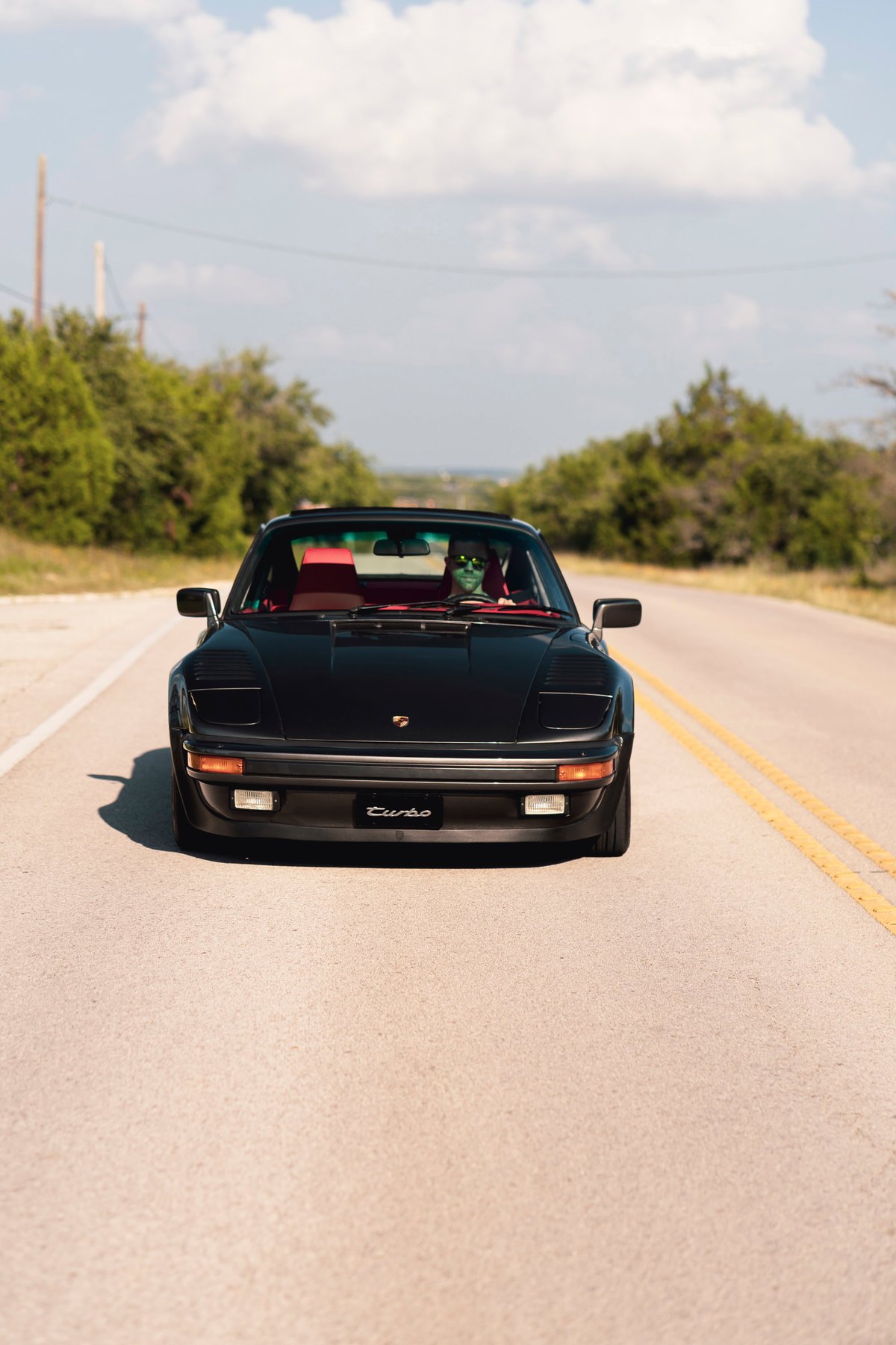 Front view of a black 1989 Porsche 930 Turbo Flachbau driving on a road. Red interior visible.