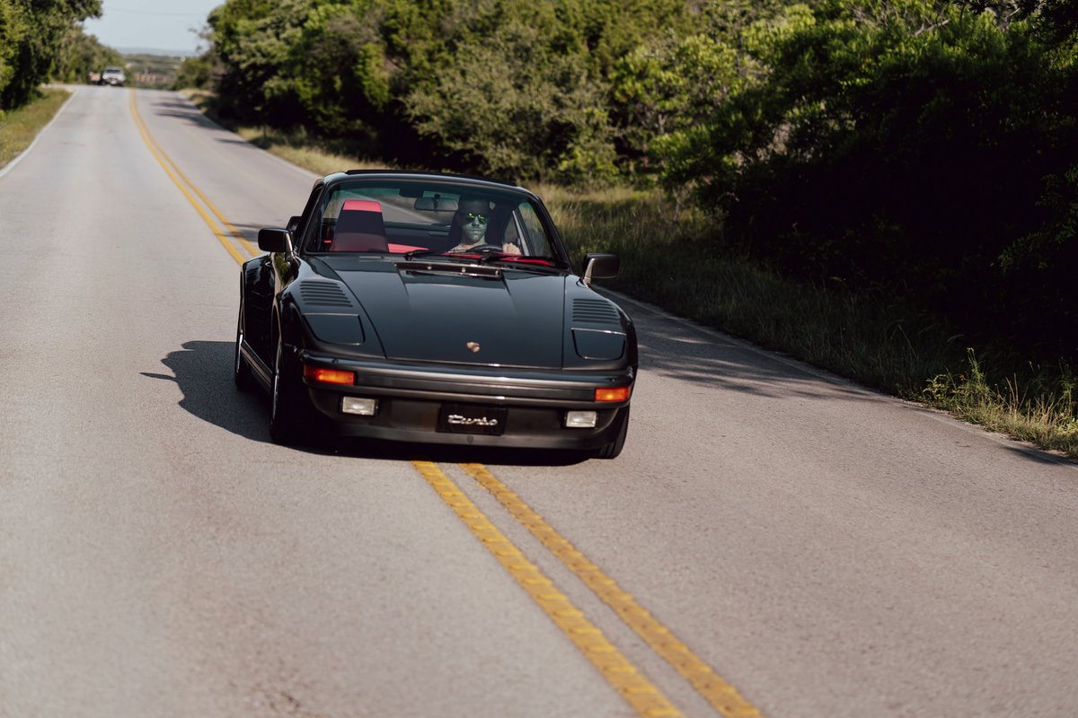 Black 1989 Porsche 930 Turbo Flachbau with red interior drives on a country road.